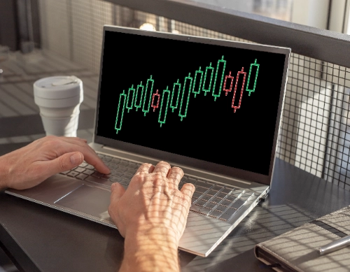 Un hombre escribe en una laptop que muestra un gráfico de velas financieras en su pantalla.