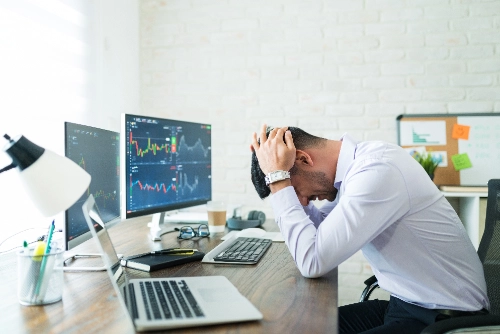 Un hombre frustrado se toma la cabeza con las manos frente a un escritorio con varios monitores que muestran gráficos financieros.