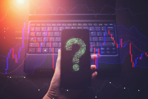Una mano sostiene un teléfono que muestra un signo de interrogación digital verde, con una laptop y gráficos financieros al fondo.