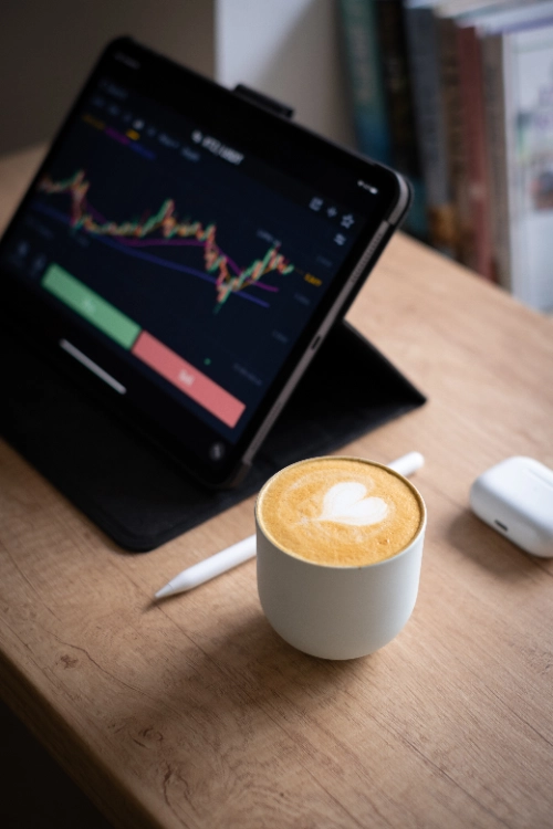 Una taza de café con arte en espuma descansa sobre un escritorio de madera junto a un lápiz óptico, un estuche de audífonos y una tableta que muestra un gráfico financiero.