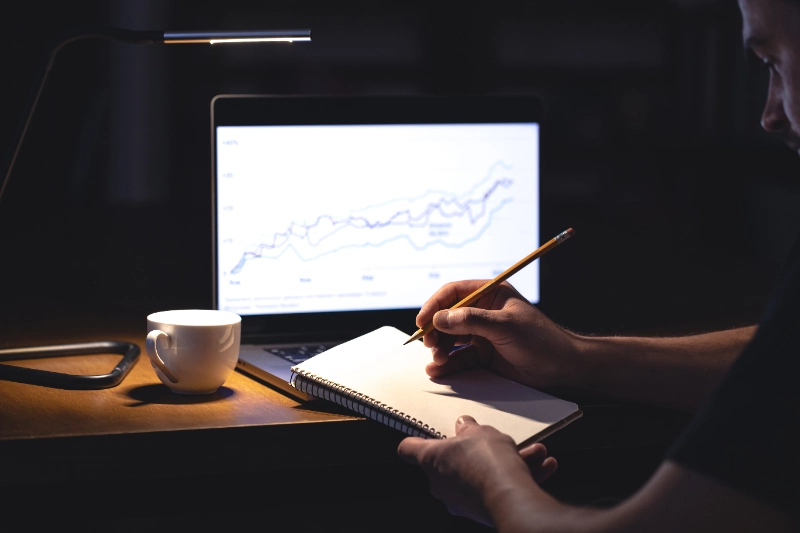 Diario de trading: hombre frente al ordenador con gráficos y una libreta por la noche