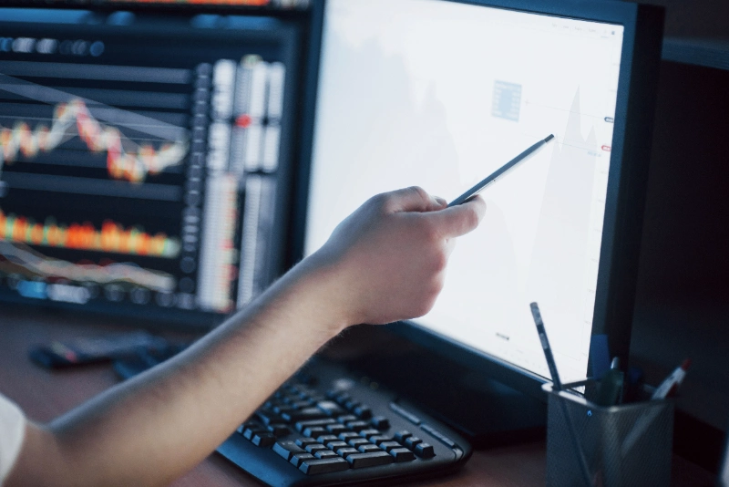 Mano de un profesional señalando un gráfico financiero en un monitor con un bolígrafo, representando el análisis técnico de datos y la supervisión del mercado bursátil.